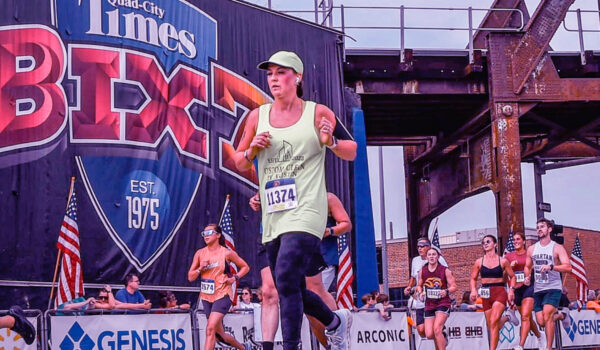 Photo of Rachel Loussaert running on asphalt in the Bix 7 marathon in Davenport, Iowa, with part of a rusted bridge, and other runners in the background.